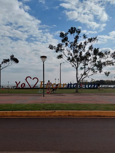In Encarnación, Paraguay, a street-level, medium shot captures a park area adjacent to a body of water, likely the Paraná River. In the background, large, three-dimensional letters spell out "Yo Amo Encarnación," with a prominent red heart symbolizing love for the city. Several people are present. Two individuals, a woman and a man, are seated on chairs facing away from the camera, near the word "Encarnación." To the left, a young boy in an orange shirt stands near the water's edge. Further back, a family or group of individuals are interacting on the grass in front of the letters, with one person appearing to hit a ball with a racket. A large tree with dark foliage dominates the right side of the frame, its branches extending across the sky. The sky itself is partly cloudy, with patches of blue visible. A lamppost stands prominently in the middle ground. The foreground consists of a paved walkway with red and beige patterned bricks, bordered by a strip of green grass and a yellow curb, leading down to a dark asphalt road.