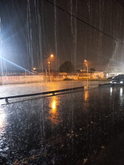 The image depicts a heavy rainfall scene at night in the city of Quevedo, Ecuador. The foreground shows wet asphalt with visible reflections of streetlights. Vertical streaks of heavy rain dominate the view. In the midground, there is a low barrier along the edge of the street, and beyond it, streetlights illuminate buildings and a wall with faded graffiti. A vehicle with its headlights on is parked further down the street. The overall environment suggests an urban setting during a severe downpour.