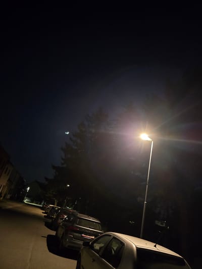 The image depicts a night scene on a street in Kostelec na Hané, Czechia. A row of parked cars lines the right side of the asphalt road, which recedes into the darkness. The street is illuminated by a tall, white lamppost positioned on the right side of the road, its bright light creating lens flares and casting shadows from the trees and vehicles. In the distance, faint lights suggest buildings and other streetlights. The sky is dark blue, with a faint, diffuse object visible in the upper left quadrant, possibly the moon or a distant light source. The overall atmosphere is quiet and still, characteristic of a residential street at night.
