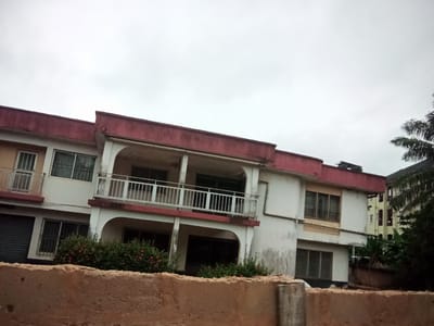 This is a medium-angle, eye-level shot of a two-story, multi-unit residential building in Nanka, Nigeria, under an overcast sky. The building features a prominent red trim along its roofline and balconies. The facade is primarily white, with some sections appearing weathered or stained.
On the left side of the building, a light yellow section with a metal roll-up door is visible on the ground floor. The first floor has a window with a barred frame. Adjacent to this, a white section of the building has two windows on the ground floor, both with barred frames, and a larger window on the first floor.
The central part of the building is characterized by a wide, covered balcony on the first floor, supported by pillars. This balcony has a white metal railing. Below the balcony, on the ground floor, there are two dark-windowed openings. A dense green shrubbery with small pink flowers is planted in front of this section.
To the right, the building continues with windows on both levels, also featuring barred frames. A lighter yellow building is visible behind this section, and further to the right, dark green foliage, likely trees, can be seen.
In the foreground, a rough, reddish-brown concrete wall runs horizontally across the bottom of the frame, partially obscuring the base of the building. The wall appears to be made of stacked blocks, with some visible texture and imperfections. The overall impression is of an urban residential area with a somewhat aged but functional building.