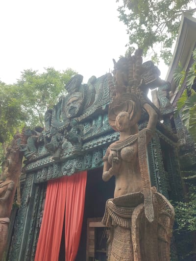 The image shows an ornately decorated temple entrance in Muang Pattaya, Thailand. The entrance is framed by intricately carved stone structures featuring mythical creatures and figures. A red curtain hangs within the entrance. To the right stands a weathered statue of a female figure, adorned with elaborate headwear and jewelry. The background includes lush greenery, suggesting a garden setting. The top of a building is visible on the right. The sky is overcast.