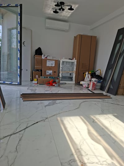 The image depicts the interior of a room under construction or renovation in Dolna Malina, Bulgaria. The floor is covered in large, white marble-patterned tiles. Sunlight streams in from an open doorway on the left, casting distinct shadows across the floor.
In the foreground, several long, dark wooden planks are laid parallel to each other. A grey cable runs diagonally across the floor from the lower left to the upper right.
The room is filled with various construction materials and appliances. On the left, a large, open doorway reveals a glimpse of greenery outside. To the right of the doorway, a tall white wardrobe or packaging stands against the wall, bearing a large stylized "E" logo and the text "1:2000".
Against the back wall, several cardboard boxes are stacked, some appearing to be appliance boxes. A black backpack is placed on top of some of these boxes. On top of the boxes, various items are visible, including what appears to be a white appliance with a grey control panel, possibly a washing machine or dryer, packaged in white foam.
Further to the right, a Husqvarna chainsaw with an orange casing rests on the floor. Next to it, a yellow and black piece of equipment, possibly a generator or a large tool, is visible. A stack of items wrapped in plastic bags, containing red and white packaging, is also present.
On the far right of the image, two dark-framed doors with frosted glass panels are propped against the wall, along with several other vertically stacked wooden planks.
An air conditioning unit is mounted high on the white wall above the boxes. A black, modern ceiling light fixture with three lamps is also visible. The walls are painted white.