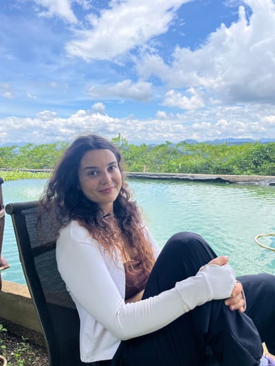 A woman with long, curly brown hair is sitting in a black chair next to a pool of turquoise water. She is wearing a brown top and a white jacket. Her hands are resting on her lap, and she is looking at the camera with a slight smile. The background shows a lush green landscape with mountains in the distance and a blue sky with white clouds. The scene appears to be outdoors in a tropical or subtropical region. The location is identified as Sơn Điền, Vietnam.