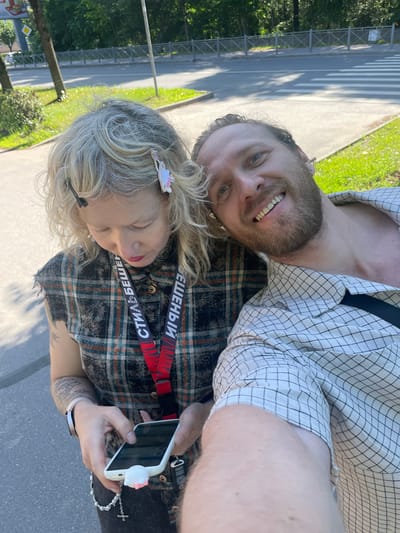 This is a candid, slightly blurry selfie taken outdoors. The camera is held by a woman with blonde, curly hair, who is looking down. She is wearing a plaid shirt and a lanyard with red and black straps that has text on it. A white phone with a pink cat-shaped attachment is visible in her hands. To her right is a man with a beard and blue eyes, smiling at the camera. He is wearing a checkered shirt. In the background, a street with a pedestrian crossing and greenery can be seen, suggesting a daytime setting on a sunny day. The overall mood appears to be relaxed and casual. The visible text on the lanyard appears to be in Cyrillic, and includes the words "БЕШЕНЫЙ" and "СТИЛЬ".