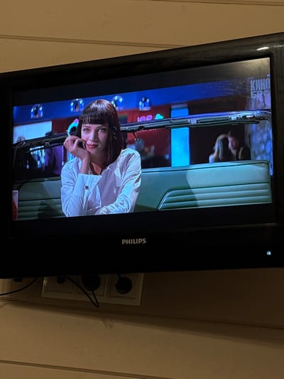 A Philips television, displaying content from the film *Pulp Fiction*, is depicted in a room within the city of Zelenogradskiy, Russia. The screen shows the character Mia Wallace, portrayed by Uma Thurman, seated in what appears to be a diner booth. She has dark, shoulder-length hair with bangs, is wearing a white long-sleeved shirt with cufflinks, and is positioned on the left side of the screen, looking towards the viewer with a slight smile, her right hand resting under her chin. The background of the film scene features blue and red neon lighting with geometric patterns, and further to the right, two blurred figures are visible, indicative of other patrons. The top right corner of the screen shows Cyrillic text, "КИНО БР," indicating a broadcasting or streaming source. The television itself has a black frame, and a small white indicator light is illuminated below the screen near the "PHILIPS" brand logo. Below the television, mounted on a light-colored wall with horizontal paneling, is a white triple electrical outlet. Two black power cords are plugged into the left and middle sockets, extending downwards.