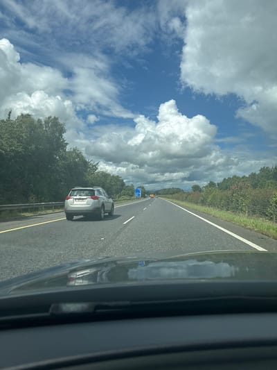 This is a landscape photograph taken from the passenger seat of a car, looking out at a highway. The image is oriented vertically, with the road and sky occupying the left and right sides of the frame, respectively. The road, with a white line marking the lane, stretches into the distance and appears to be a multi-lane highway. A silver SUV is visible in the right lane, and further ahead, another car and a blue sign are discernible.
The sky is predominantly blue with large, fluffy white clouds, indicating fair weather. Trees line the left side of the highway, forming a dense green barrier. The right side of the frame is dominated by the reflection of the sky and clouds on the dark, glossy surface of the car's exterior. A metal guardrail is visible along the side of the road.
The time of day appears to be daytime, with the sun illuminating the scene. The overall impression is that of a typical road trip or commute on a pleasant day. No people are clearly visible. The license plate of the silver SUV reads "EZ JNH". There is a blue highway sign visible in the distance with what appears to be white text and symbols.