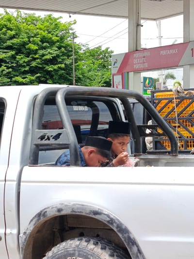 Two young boys wearing black peci caps are in the back of a white pickup truck, possibly at a gas station in Cotbadatunong, Indonesia. One boy is eating from a plastic container, while the other looks towards the camera. The truck has a roll cage and "4x4" branding. In the background, a gas station sign displays "PERTAMINA" and fuel types. Yellow crates are stacked behind the truck. Lush green trees are visible to the left, and a cloudy sky is overhead. The scene appears to be during daylight hours.