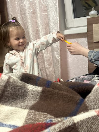 A young child lies on a patterned surface, possibly a bed, with a hand reaching towards her, offering a yellow object. The child is wearing a patterned jumpsuit, and her hair is adorned with a purple flower clip. A blanket with a plaid pattern is partially obscuring the view, and a window with a baby bottle attached to the frame is visible in the background. The location is Uva, Russia.