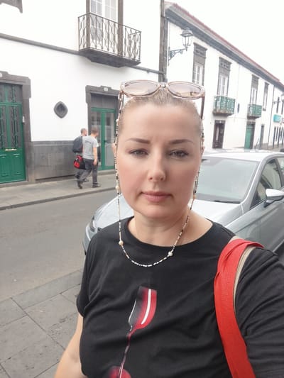 A woman takes a selfie outdoors, wearing sunglasses on her head and a black t-shirt with a red and white graphic. She has a beaded necklace around her neck. Behind her, a silver car is parked on the street. Further in the background, two men walk down a sidewalk in front of a white building with green doors and balconies. The scene appears to be a street in Ribeira Grande (Conceição), Portugal. The sky is overcast, suggesting a cloudy day. The woman's expression is neutral.