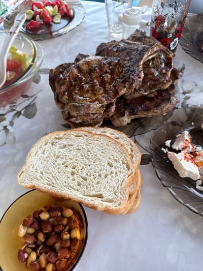 A close-up, eye-level shot captures a rustic meal spread on a white tablecloth with a floral pattern, possibly in Ravadinovo, Bulgaria. The main subject is a stack of grilled meat, likely steaks, with visible char marks and a glistening, browned exterior. To the left of the meat are slices of white bread, their porous texture evident. A small, amber-colored bowl filled with roasted peanuts sits in the foreground, partially obscured by the bread. To the right of the meat, a dark, wavy-edged plate holds a white cheese, likely feta or a similar soft cheese, sprinkled with red spice, possibly paprika. In the background, two glass dishes containing chopped tomatoes and cucumbers, dressed in a dark liquid, suggest a salad or appetizer. A glass with a decorative floral print, possibly holding a beverage, and a salt shaker are also visible in the background, hinting at a meal setting. The lighting is natural and diffused, creating soft shadows and highlighting the textures of the food. The overall impression is one of a hearty, casual outdoor meal.