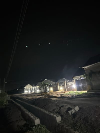 A nighttime scene shows a building illuminated by lights, adjacent to a steep, dirt slope reinforced with concrete. Power lines cut across the lower left of the frame, and a dark, undefined area occupies the majority of the left side. Small, indistinct green lights appear in the dark space. The setting has a gritty, somewhat desolate appearance.