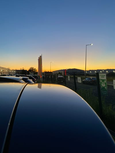 The image depicts a sunset scene in Bolesławiec, Poland. In the foreground, the glossy, dark surface of a car roof reflects the sky and surrounding objects. The middle ground features a parking lot with several cars and a chain-link fence. Beyond the fence, industrial buildings and a streetlamp are visible, illuminated by the warm glow of the setting sun. The sky transitions from a deep blue at the top to a vibrant orange and yellow near the horizon. There are no people visible in the image, and the overall atmosphere is one of quiet twilight.