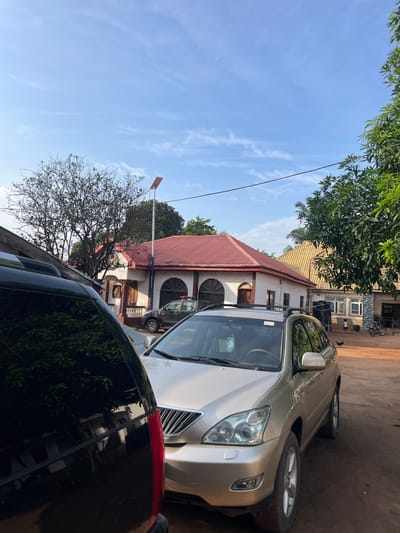 The image depicts a daytime scene in the city of Amurie, Nigeria. In the foreground, a portion of a black SUV is visible on the left, with its reflection of trees and buildings on its polished surface. To the right of the SUV is a silver SUV, facing towards the right, with its front end and driver's side visible. Behind the SUVs, a building with a red tiled roof and white walls is prominent, featuring arched windows and decorative columns. To the right of this building, another structure with a yellowish, sloped roof is visible, with a dark water tank against its wall. The ground is a reddish-brown dirt. A street light is mounted on a pole in front of the building with the red roof. The sky above is a clear blue with scattered white clouds. In the background, there are trees and what appears to be a police vehicle parked near the red-roofed building. A few chickens are present on the ground in the middle distance.