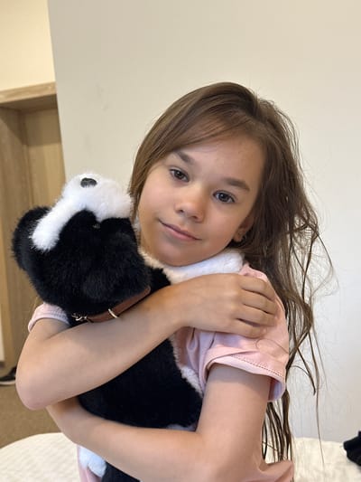 A young girl with long, dark brown hair is lying down and holding a stuffed animal that resembles a skunk. She is wearing a light pink shirt. The stuffed animal has black and white fur and appears to be a toy. The girl is looking towards the camera with a gentle smile. Her arms are wrapped around the stuffed animal. The background is a plain, light-colored wall, suggesting an indoor setting. The lighting is soft and natural. There is no visible text in the image. The overall mood is one of comfort and affection towards the toy.