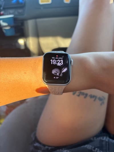 A close-up, vertical shot shows a person's arm wearing a silver Apple Watch with a light pink band. The watch face displays "FRI, 26 JUN", the time "19:23", a stylized graphic resembling a face, and the temperature "28°". The band has a repeating pattern of outlined heart shapes.
The person's skin appears tanned and lightly freckled. A faint scar is visible on the forearm above the watch. To the right of the arm, a tattoo with cursive text, possibly "You love Self", is partially visible on the upper arm or shoulder. The background is blurred, but suggests an indoor setting with furniture and reflective surfaces, possibly a room or a vehicle interior. Sunlight casts a warm glow on the arm. The time displayed on the watch indicates it is late afternoon or early evening.