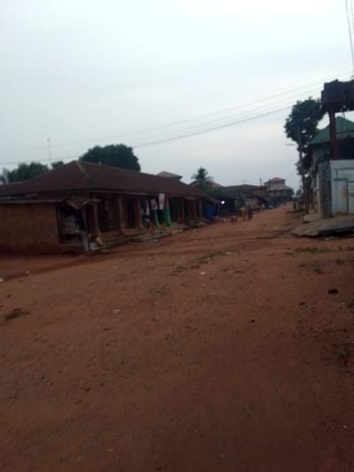 This image captures a street scene in what appears to be a rural or semi-urban area, consistent with Orlu, Nigeria. The dominant feature is a wide, unpaved road made of reddish-brown dirt or laterite, stretching into the distance. The road surface shows variations in texture and some small patches of sparse greenery.
On the left side of the road, a long, low-slung building with a dark, corrugated metal roof and what appears to be unpainted earth or cement walls stands prominently. It features multiple open doorways or shopfronts along its facade. Colorful fabrics or clothing are seen hanging outside one of these openings, suggesting a market or retail activity. Further down the street on the left, other buildings and structures, some partially obscured, continue to line the road.
On the right side of the road, closer to the foreground, a building with contrasting lighter-colored walls and a textured, possibly stone-clad section is visible. Above it, a dark, elevated structure, potentially a water tank stand or a billboard frame, is silhouetted against the sky. Power lines or utility wires traverse the upper part of the frame.
The sky is uniformly overcast and light grey, casting a dim, diffused light over the entire scene. This suggests either early morning, late afternoon, or a heavily clouded day. A few indistinct figures of people can be seen further down the road, walking or standing, indicating some daily activity. Trees and foliage are scattered throughout the background, adding to the natural setting. There is no discernible text visible in the image.