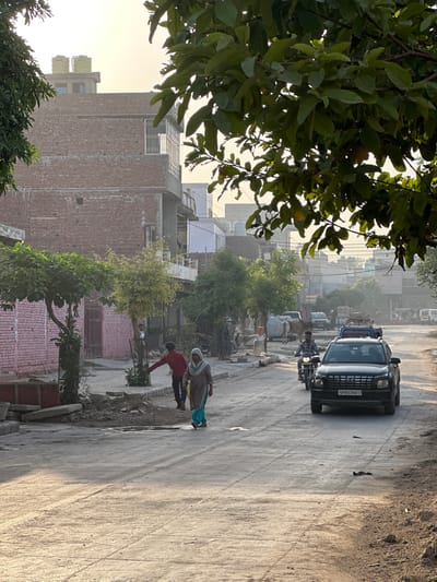 Captured in Mathura, India, the image portrays a street scene during daylight. A black car with the license plate "UP85CV0017" moves along the street, followed by a person on a scooter. In the middle ground, two individuals are near the edge of the road; one in a red shirt is bent over, possibly tending to plants, while the other, wearing a headscarf, walks nearby. Along the street, various vehicles, including trucks, and a person with a cow are visible. Brick buildings line the road, with lush foliage in the foreground and background framing the view.