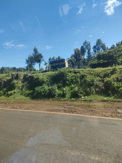 The image shows a view from a vehicle driving along a paved road. The sky is bright blue with a few scattered clouds. Power lines stretch across the upper portion of the image. To the side of the road is a raised embankment covered in lush green vegetation, including grass, bushes, and trees. On top of the embankment, there is a small structure with blue and white signage, surrounded by a metal fence. The location appears to be a rural area in Kenya. The Municipality ward is unknown. No people or interactions are visible.