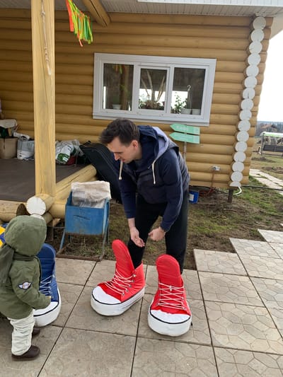 In Sepych, Russia, a man stands outdoors wearing oversized red and white sneaker costumes, bending over a blue metal box, possibly examining or using it. He is dressed in a dark jacket and pants. To his right, a child is sitting with their back facing the camera, clad in an olive green jacket and beige pants, with similarly oversized blue and white sneaker costumes on their feet. They are situated on a tiled surface. Behind the man is a wooden structure with a window and decorative elements, possibly a building or cabin. The environment includes vegetation and an overcast sky. Several bags and other items are visible near the building's base.