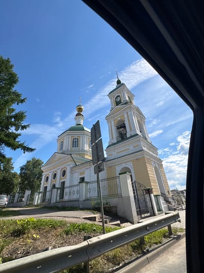 This media file captures a view of a church, likely in Petrovskoye, Russia. The church has a distinct architectural style, featuring a tall bell tower with a clock and a smaller dome topped with a cross. The building itself is painted in a light yellow with white trim and has several arched windows and decorative elements. It appears to be situated in a somewhat urban or suburban setting, with a road and grassy area visible in the foreground, separated by a metal guardrail. Trees are also present, suggesting a location with greenery.

The image is taken from a low angle, looking upwards towards the sky, which is a clear blue with scattered white clouds. This indicates fair weather. The lighting suggests it is daytime, likely midday or afternoon, given the brightness and shadows. There are no visible people in the frame, and the primary focus is on the church and its surroundings. The scene conveys a sense of tranquility and architectural beauty. No visible text is present in the image.