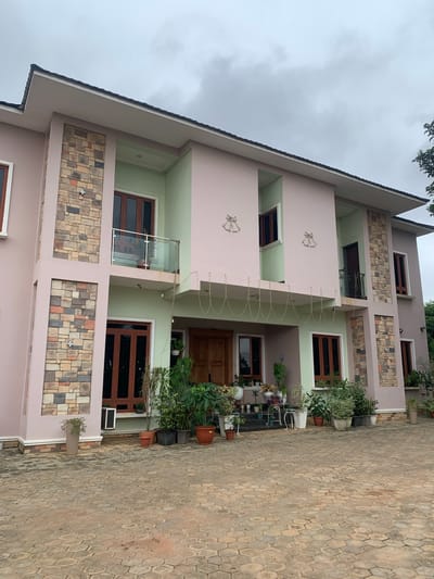 The image depicts a two-story house with a light pink and mint green exterior, located on a cobblestone driveway in Onibudo, Nigeria. The house features prominent stone accent walls on the facade and multiple windows with dark wood frames. A glass-railed balcony is visible on the upper floor, and decorative bell shapes are mounted on the upper section of the house. The entrance area is adorned with a variety of potted plants and greenery, suggesting a well-maintained garden. The sky is overcast, indicating a cloudy day. No people are visible in the image.