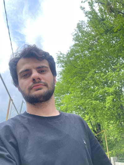 The image shows a man with dark hair and a beard wearing a black shirt. He appears to be outdoors, likely in Chiatura, Georgia, as suggested by the presence of distinctive cable car infrastructure often found in the area. The background includes green trees and a partly cloudy sky.