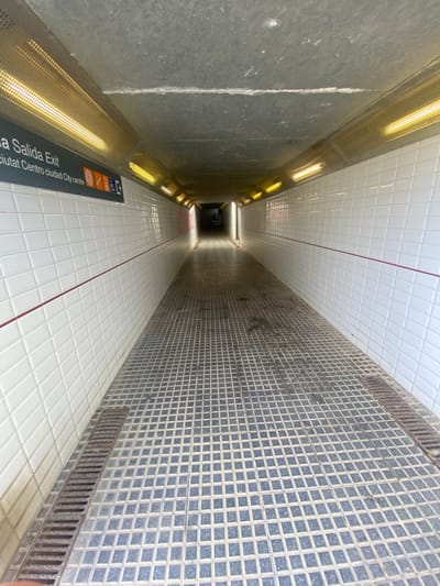 This is a forward-facing, eye-level shot of a pedestrian underpass in Vilassar de Mar, Spain. The underpass is brightly lit by rows of yellow fluorescent lights recessed into the ceiling on either side. The walls are covered in white rectangular tiles, with a thin red line running horizontally at eye level down the center of each wall. The floor is paved with dark, square, textured tiles, forming a grid pattern. On the left wall, a dark sign reads "Salida Exit" in white text, followed by pictograms for a circular target, a wheelchair accessible symbol, and an arrow pointing forward. The underpass recedes into the distance, with a person visible at the far end. The overall impression is one of clean, functional, urban infrastructure.
