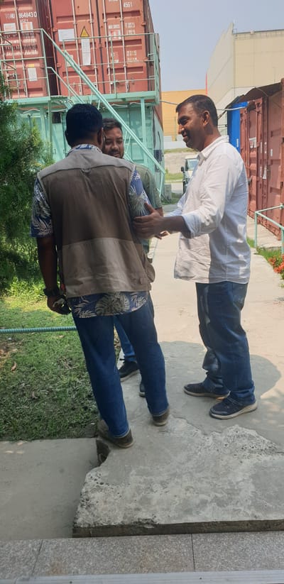 In Rooppur, Bangladesh, three men are engaged in a conversation outdoors. The man on the left, viewed from behind, wears a patterned blue and white short-sleeved shirt with a khaki vest over it and blue jeans. He holds a dark object in his right hand. The man in the middle, facing left, is wearing a green shirt and blue jeans. The man on the right, facing left and slightly smiling, wears a white long-sleeved shirt and blue jeans. He is gesturing with his hands as he speaks to the other two men. The setting appears to be an industrial or construction area, with stacked shipping containers, including one labeled "45G1", visible in the background. A tree with green foliage is on the left, and a paved pathway with some cracked concrete is in the foreground. The sky is clear and bright.