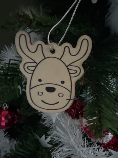 A close-up image captures a light brown, flat, reindeer-shaped ornament with black outlines defining its face, including two black dot eyes, a larger black oval nose, a smiling mouth, and stylized antlers. A thin, silver-colored string forms a loop through a small hole at the top of the ornament, suspending it. The ornament hangs on an artificial Christmas tree, characterized by dark green pine needles visible in the background. Other decorations partially observed include a shiny red spherical ornament in the lower-left foreground and segments of white and red tinsel or garland in the lower-right background. The scene is located in the city of Le Puy-en-Velay, France.