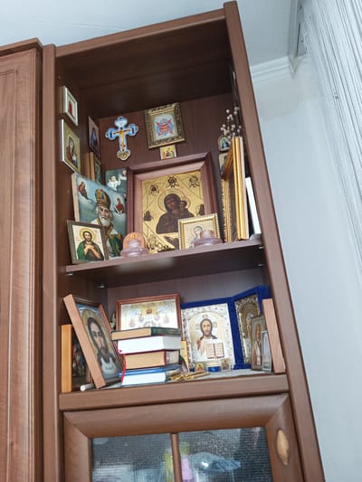 The image displays a tall, dark brown wooden cabinet with several shelves filled with religious icons and other devotional items. The cabinet is positioned against a white wall, with a white string curtain visible on the right side of the frame. The items on the shelves include framed icons of religious figures, a blue and gold cross, small statues, and a stack of books. The overall arrangement suggests a home altar or a dedicated space for religious veneration. The lighting is consistent with indoor ambient light. The location is identified as Maryanskaya, Russia.
