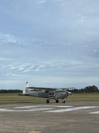 A single high-wing, propeller-driven aircraft, bearing the registration "LV-CQS" on its vertical stabilizer, is stationary on a paved runway or taxiway in Chascomús, Argentina. The aircraft is primarily dull grey with a green engine cowling, and its pilot-side door is visibly ajar. White painted numerals or letters are partially visible on the concrete surface in the foreground. The immediate environment comprises a flat, green grassy field, which transitions into a dense line of dark green trees along the horizon. The sky is uniformly overcast with light grey clouds, suggesting diffuse daylight conditions. No individuals are present within the visible scene.