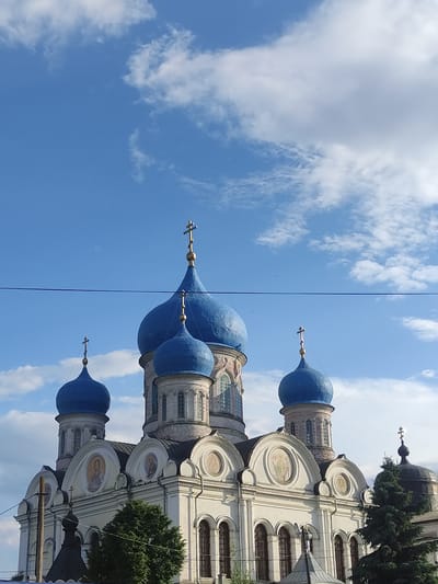 This image shows a medium shot of the Church of the Transfiguration, a domed Orthodox church located in Rogachovo, Russia. The church has a white facade with several blue domes topped with golden crosses. The main dome is the largest, with smaller domes surrounding it. The church features arched windows and decorative elements, including circular paintings on its facade. In the foreground, a power line runs horizontally across the frame. Green trees are visible to the right and left of the church, and a dark, smaller dome structure is partially visible on the right. The sky above is blue with scattered white clouds. No people or actions are depicted in the image.