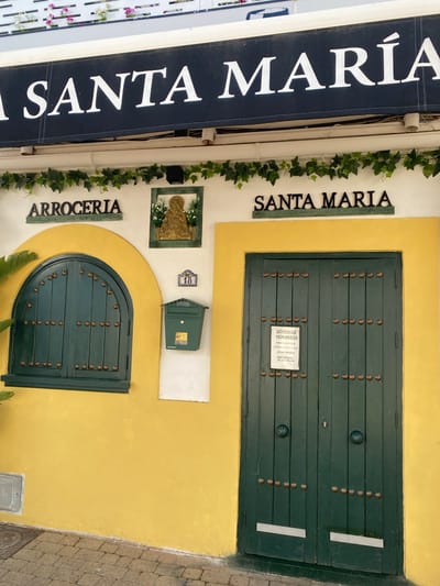 A medium shot captures the exterior of a building in Cancelada, Spain, featuring a prominent awning with white lettering that reads "A SANTA MARIA." Below the awning, a green garland of ivy adorns the upper facade. To the left, a sign in dark lettering on a green background states "ARROCERIA," indicating a rice restaurant. Adjacent to this sign, a small framed relief depicts a religious figure with outstretched hands, adorned with white flowers. A dark green, arched window with studded detailing is to the left of the relief. To the right of the window and below the relief, a green mailbox is mounted on the wall, with a small plaque reading "18" above it. To the right of the mailbox, a large, dark green double door with similar studded detailing dominates the lower section of the facade. A white sign is attached to the right door, displaying operating hours and reservation information. The building's lower section is painted a vibrant yellow, contrasting with the white upper walls. The ground in front of the building is paved with cobblestones.