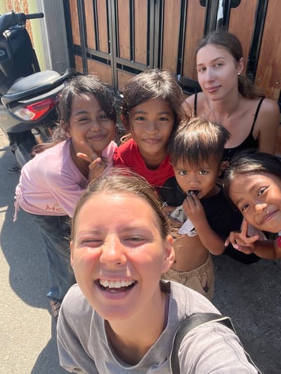 In Kertasari, Indonesia, a group of five children and two adults pose for a selfie outdoors. The children, with varying skin tones and dark hair, are arranged in front of a wooden gate. One young girl wears a pink t-shirt, another a red t-shirt, and a young boy is dressed in a black t-shirt. The other two children are partially visible. Two adults are also present, one with light brown hair and a black tank top, the other with blonde hair and a gray t-shirt, with her face filling the foreground of the image. A motorcycle is parked to the left of the group. The ground is paved with asphalt.