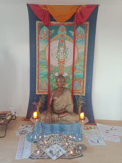 The image depicts an indoor Buddhist altar. The focal point is a wooden Buddha statue seated in meditation on a blue cloth-covered platform. Behind the statue is a large thangka painting with various figures and landscape elements, framed by a dark blue border. The thangka is topped with draped fabrics in red and orange.
On either side of the statue, there are candles and small vases with greenery. In front of the altar, scattered on the floor, are several lit candles, a patterned cloth, and various drawings on paper, likely created by children. A low table with books and other objects is visible on the left side of the frame.
The room has light-colored walls and a wooden floor. The overall impression is one of a personal and lovingly assembled space for meditation and spiritual practice. The image was taken in Jérica, Spain.
