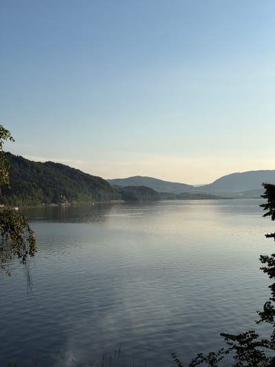 The image depicts a serene lake scene with rolling hills covered in lush green trees in the background. The sky is a clear, pale blue, transitioning to a lighter hue near the horizon, suggesting it is either early morning or late afternoon. The lake's surface is calm, with gentle ripples reflecting the soft light.
In the mid-ground, a densely forested hill slopes down towards the water's edge. A small cluster of buildings, appearing to be houses or a small village, is nestled on the hillside amongst the trees. There are a few boats visible near the shore, indicating potential recreational activity.
Further in the distance, more hills and mountains can be seen, some shrouded in a light haze, giving the scene a sense of depth. The overall impression is one of natural beauty and tranquility, with no visible people or immediate action taking place. The location is identified as the city of Lipie, Poland.