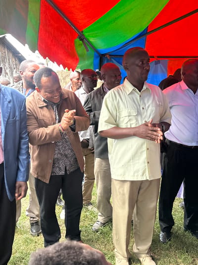 A group of men is gathered outdoors under a vibrant red, green, and blue striped tent in Kiganjo, Kenya. The image, captured in bright daylight, shows the men standing in rows, looking forward and slightly upwards, suggesting they are attentive to a speaker or a focal point outside the frame.

In the foreground, a prominent man in a light yellow collared shirt and khaki trousers stands with his hands clasped in front of him. To his left, another man in a brown jacket over a patterned shirt also stands with clasped hands, sporting a colorful beaded bracelet on his left wrist. Other men are visible behind them, dressed in a mix of collared shirts, jackets, and trousers in various neutral tones like white, grey, and black. Some wear glasses. Their attire ranges from semi-formal to casual, appropriate for an outdoor gathering or community event.

The ground beneath them is covered in green grass, indicating an outdoor setting. The colorful tent panels provide shade, suggesting sunny weather. The men appear calm and engaged, with no strong emotions overtly displayed. Their posture implies respect and attentiveness. Partially visible in the far left background is a wooden structure, possibly a fence or part of a building, hinting at the local environment. There is no visible text in the image.