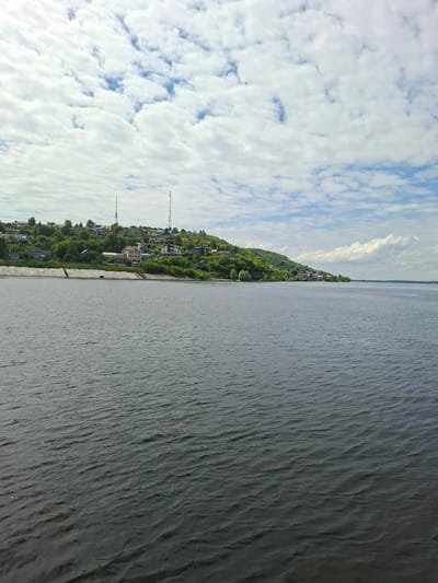 This is a landscape image captured from a side-on perspective, divided into two distinct halves: the sky and the water. The left half of the image is dominated by a cloudy blue sky. Fluffy white clouds are scattered across the expanse, with patches of blue sky visible between them. In the distance, along the horizon, a thin line of land is visible.
The right half of the image is a body of water, likely a river or lake, with a textured surface. The water is dark and appears to have ripples or small waves, suggesting a gentle breeze.
In the middle ground, along the shoreline, is a settlement situated on a gentle slope. Buildings with varying roof colors, interspersed with lush green trees, are visible, suggesting a populated area nestled amongst nature. Two tall communication towers are also present, breaking the natural skyline. The shoreline itself is lined with a concrete embankment.
The image appears to have been taken during the daytime, with the lighting suggesting it could be late morning or early afternoon. The presence of clouds indicates a partly cloudy day. The scene is tranquil and offers a view of a semi-rural or suburban setting by a large body of water. No text is visible in the image. The orientation of the image suggests it might have been taken with the camera held vertically.