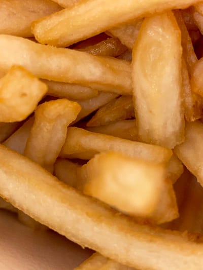 The image presents a close-up, top-down perspective of a dense pile of golden-brown french fries. The fries are numerous, elongated, and irregularly shaped, filling the entire frame. Their surfaces display varying shades, ranging from light golden to deeper caramel brown, indicative of frying. Some pieces exhibit a slightly crisp or textured exterior, while others appear smoother. The individual fries are arranged haphazardly, overlapping and interlocking. They appear to be contained within an indistinct, light-colored vessel, visible only as soft edges along the bottom and left sides of the frame. No people, specific actions, or identifiable interactions are visible. The image contains no visual elements or contextual information that would allow for the identification of a specific location within Seeb Province, Oman, or any other geographical area.