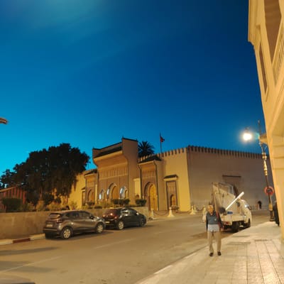 The image shows a street scene in Mechouar Fes Jdid, Morocco, at night. The sky is a deep blue. On the left, a gray Renault Captur and a dark car are parked along the street. A building with decorative arches and a crenellated wall is visible in the background, adorned with a Moroccan flag. A palm tree stands behind the building. A man walks on the sidewalk in the foreground, wearing light trousers, a blue and gray shirt, and carrying a red bag. Behind him, a white truck is parked. A streetlight illuminates the scene on the right side. Part of a building facade appears on the right. Another person is walking further down the sidewalk near the truck.