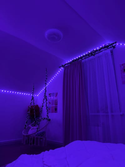 The image depicts a bedroom illuminated by purple LED strip lights, creating a moody ambiance. The lights are installed along the ceiling and around the window area, with artificial greenery intertwined with the lights above the curtain rod. A pink curtain and sheer white curtains cover a window, behind which a radiator is visible. In the corner of the room, a white macrame hanging chair with a stuffed animal is suspended. A large red rose decoration hangs above the chair. On the wall near the chair, several posters are displayed. The foreground shows a portion of a white duvet or comforter. The room appears to be a personal living space, possibly a bedroom. The location is stated as Obninsk, Russia.