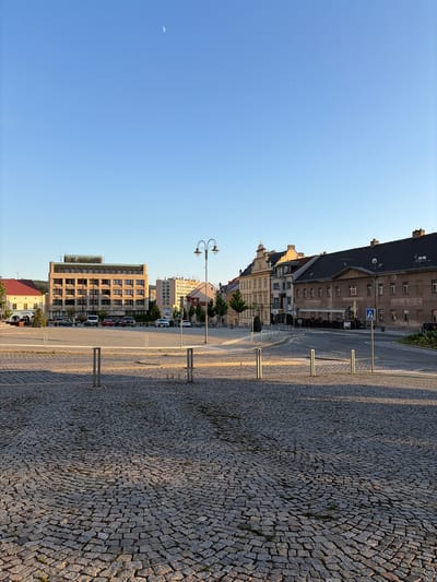 The image captures a view of a cobblestone plaza in Příbram, Czechia. In the background, a large, modern building with numerous windows stands prominently, flanked by other buildings exhibiting varied architectural styles. A street with parked cars and a traffic light is visible, along with a decorative streetlamp. The sky is clear and blue, with a sliver of the moon visible. The foreground is dominated by the textured cobblestone paving of the plaza, with metal bollards marking off sections of the space. The lighting suggests it is either early morning or late afternoon, casting shadows across the plaza.