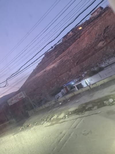 A tilted view presents a street scene with a rocky hillside to the left and a paved road to the right. Several buildings are nestled between the hillside and road. Numerous power lines stretch across the sky above. A sign stands near the bottom of the image along the road. The street appears damp or wet.