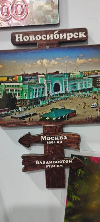 This is a close-up, low-angle, eye-level, indoor photograph of a souvenir refrigerator magnet. The magnet features a depiction of the Novosibirsk train station in Novosibirsk, Russia. Below the train station image are two wooden signposts, each shaped like an arrow. The upper signpost reads "Москва" (Moscow) with "3394 км" (3394 km) underneath. The lower signpost reads "Владивосток" (Vladivostok) with "5792 км" (5792 km) underneath. The overall scene is a still life arrangement, with no people visible. The lighting appears to be artificial, and the background is plain white. The image does not contain any information or visual cues that would place it in the city of Pokosnoye, Russia.