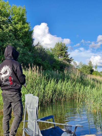 The image depicts a person standing by the water's edge, facing away from the camera, engaged in fishing. They are wearing a black hooded sweatshirt with a white dragon design and red lettering on the back, paired with dark pants. A fishing rod is visible in their hands, extending towards the water. In the foreground, two folding chairs are set up near the water, one a gray camp chair and another with a blue seat. The setting appears to be a natural environment with dense green reeds growing along the shoreline, which is partially submerged. The water is calm, reflecting the blue sky dotted with white clouds and the surrounding greenery. The location is identified as Gargždai, Lithuania. The overall scene suggests a peaceful outdoor activity.