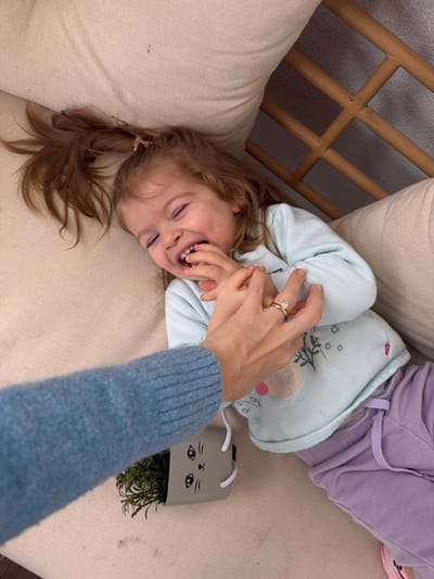 An overhead view captures a young child lying supine on a light beige upholstered surface, engaged with an adult's right hand. The child, with reddish-brown hair in two pigtails secured by pink ties, wears a light blue long-sleeved shirt featuring a floral and ice cream cone graphic with "SWEET DREAMS" text, and light purple sweatpants. The child's eyes are closed, mouth open in a wide smile, and their right hand is grasped by an adult's right hand. The adult's lower forearm, clad in a light blue knitted sweater, is visible from the lower-left. A gold ring with a clear stone is present on the adult's ring finger. Adjacent to the adult's arm on the cushion, a gray rectangular planter shaped like a cat's head with white string handles holds a small green plant. A segment of light brown wooden lattice-patterned furniture is discernible in the upper right background. The location is the city of Harish, Israel.