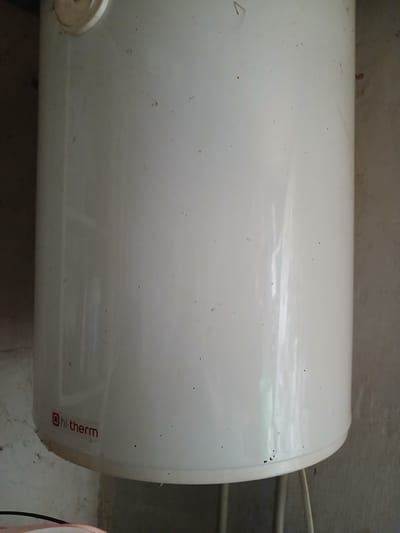 The image shows a close-up of a white, cylindrical water heater, branded with the logo "hi-therm" in red lettering. The water heater is positioned in an indoor setting with a textured, light-colored wall in the background. There are some small black specks, possibly dirt or dust, visible on the surface of the water heater. The bottom of the water heater is visible, with what appear to be pipes or supports extending downwards. The overall impression is that of a functional appliance in a utilitarian space. The location is specified as Hlushky, Ukraine.