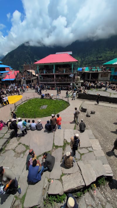 This is a high-angle, vertical, outdoor, daylight photograph taken in Malana, India. The scene depicts a village square with numerous people gathered. In the background, a prominent building with a bright red, sloped roof stands, featuring multiple stories with balconies occupied by onlookers. To the left, other buildings with red and blue roofs are visible, and further back, a verdant mountain range is shrouded in dramatic, cloudy skies.
The foreground and midground are filled with people sitting and standing on stone steps and the paved ground. Many individuals are wearing traditional clothing, including caps. In the center of the square, a circular area is covered in lush green grass. A large yellow tarp is spread out on the ground to the left of the grassy area, near a group of standing people. The ground is paved with large, irregular stone slabs, with patches of grass growing in between. Scattered throughout the scene are various individuals engaged in conversation or observing the surroundings. The overall atmosphere suggests a community gathering or event.