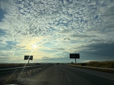 The image depicts a highway in Cherna Gora, Bulgaria, under a cloudy sky with the sun partially visible. The highway stretches into the distance, with guardrails lining both sides. On the right side of the road, a billboard with text and images is visible. The landscape surrounding the highway appears to be rural, with dry grass and some distant hills. The sun's rays create a lens flare effect across the image. There are no people or vehicles clearly discernible in the foreground or midground. The scene appears to be captured from inside a vehicle, looking forward.