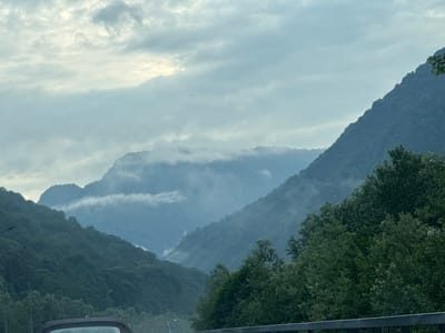 The image shows a mountainous landscape under a cloudy sky. The mountains are covered in dense green vegetation, and there are wisps of fog or clouds clinging to their slopes. In the foreground, the top of a car is visible, suggesting the photo was taken from a vehicle. The scene appears to be a view from a road, possibly a highway, given the presence of what looks like a guardrail and streetlights. The overall atmosphere is serene and slightly overcast, typical of a temperate mountainous region. This view is consistent with the scenery found in and around Chvizhepse, Russia, a location known for its mountainous terrain and natural beauty.